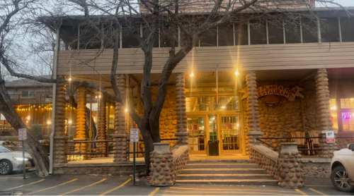 A rustic restaurant entrance with stone pillars, warm lighting, and trees in front, set in an evening atmosphere.