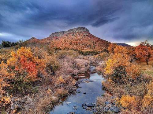 A scenic landscape featuring a mountain with autumn foliage and a stream reflecting the colorful trees under a cloudy sky.