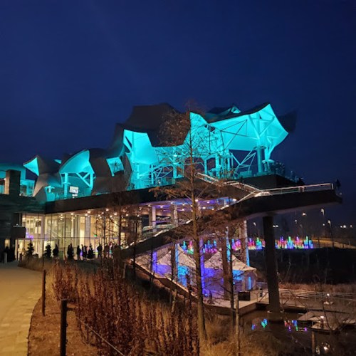 A modern building illuminated in blue and green lights at night, surrounded by trees and colorful light displays.