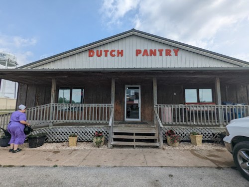 A woman in a purple dress approaches the Dutch Pantry, a rustic building with a porch and flower pots.
