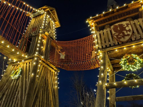 A wooden play structure with festive lights and wreaths, set against a dark evening sky.