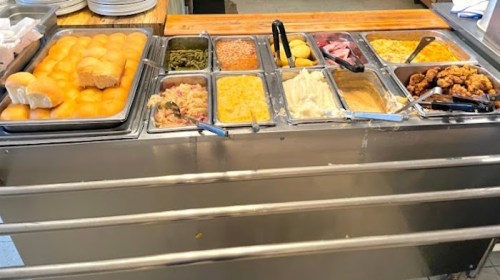 A buffet table with various dishes, including rolls, greens, rice, fruit, and fried chicken in metal trays.