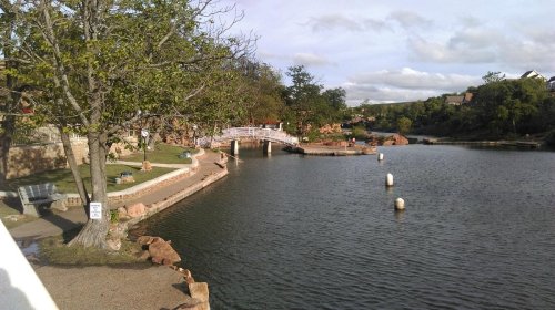 A serene park scene featuring a calm lake, a wooden bridge, and trees along the shoreline under a partly cloudy sky.