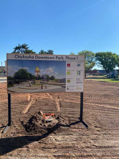 Sign for Chickasha Downtown Park Phase 1, featuring an artist's rendering and logos of project partners, on a cleared site.