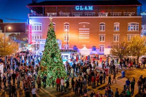 A festive crowd gathers around a decorated Christmas tree in a lively urban setting, with a building lit up in the background.