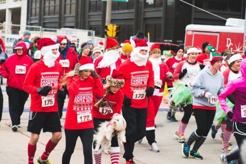 Participants in festive Santa costumes run in a holiday race, some with dogs, in a lively urban setting.