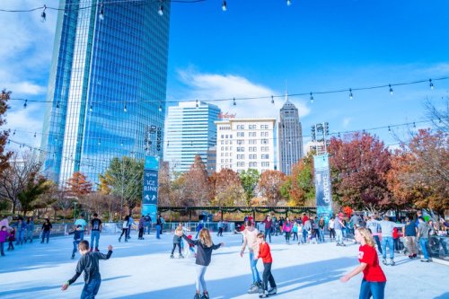 A lively ice skating rink with people skating, surrounded by colorful autumn trees and tall city buildings under a blue sky.