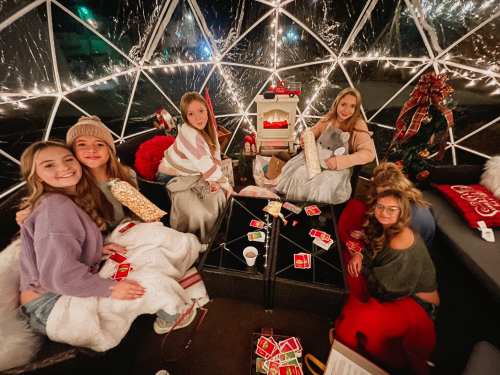 A group of five friends in cozy attire, sitting inside a festive igloo with holiday decorations and warm lighting.