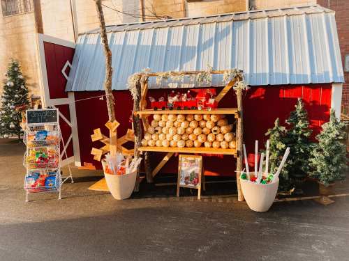 A festive outdoor display featuring a red barn, stacked baked goods, and holiday decorations with trees nearby.