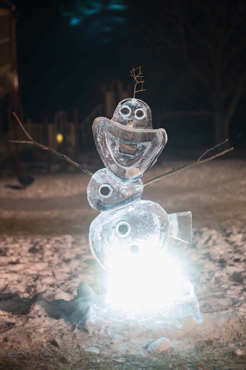 A cheerful ice snowman with a smiling face, stick arms, and a glowing base, set against a snowy background at night.
