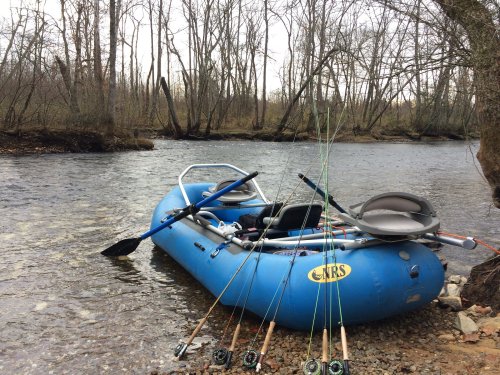 A blue inflatable raft by a river, with fishing rods leaning against it and bare trees in the background.