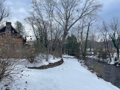 A snowy landscape with a winding path beside a stream, framed by bare trees and a stone building in the background.