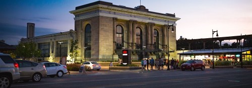Historic train station at dusk, with people gathered outside and cars parked along the street.