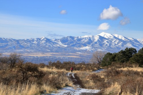 A person walks along a snowy trail with mountains in the background under a clear blue sky.