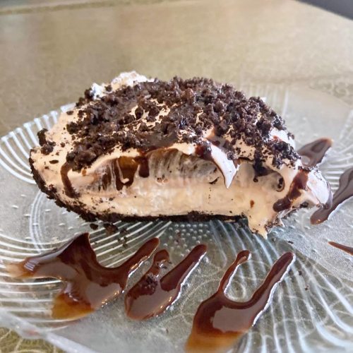 A slice of dessert with a creamy filling, topped with crushed cookies and drizzled with chocolate sauce on a glass plate.