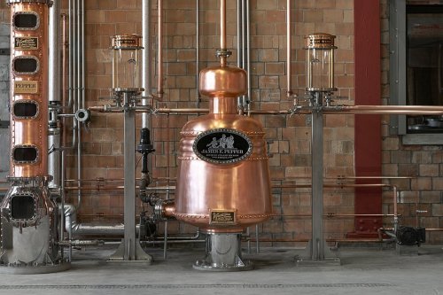 A copper distillation apparatus with metal pipes in a brick-walled distillery setting.
