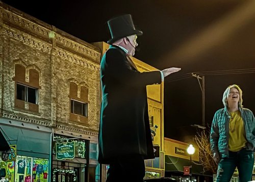 A performer in a top hat gestures while entertaining a laughing woman on a lively street at night.