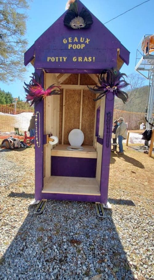 A colorful purple portable toilet decorated for Mardi Gras, with the sign "Geaux Poop Potty Gras!" and festive embellishments.