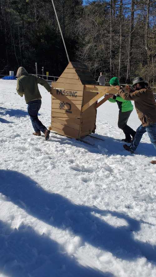 Three people pull a wooden structure labeled "PASSING #2" across a snowy landscape. Trees are visible in the background.