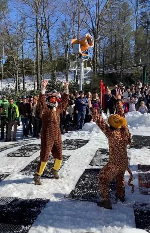 Two people in animal costumes celebrate in the snow, surrounded by a cheering crowd and snowy trees.