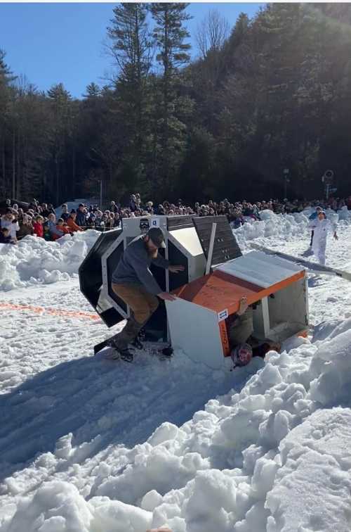 A crowd watches as a person struggles to right a flipped sled in a snowy landscape during a winter event.