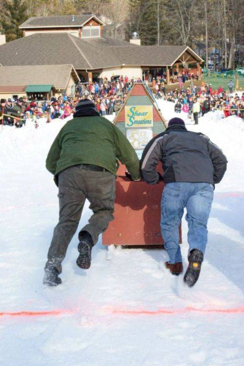 Two men push a sled down a snowy track, with a large crowd watching in the background.