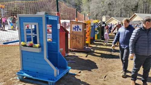 Colorful wooden structures resembling small houses line a snowy path, with people walking nearby in a winter setting.