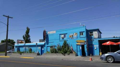 A bright blue building with signage, surrounded by greenery and parked cars, under a clear blue sky.