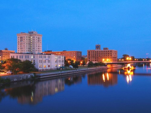 City skyline at dusk, reflecting on a calm river with illuminated buildings and a bridge in the background.