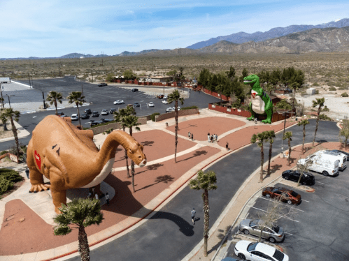 Two large dinosaur sculptures, one brown and one green, stand in a parking lot surrounded by palm trees and mountains.