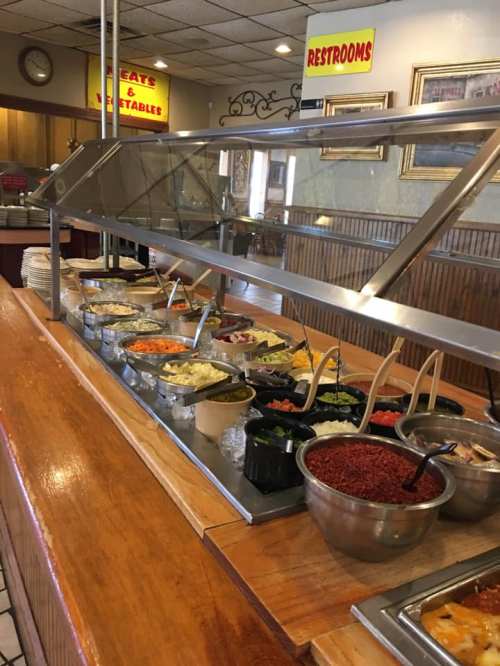 A buffet with various dishes in metal trays, including vegetables, sauces, and sides, under a glass cover.