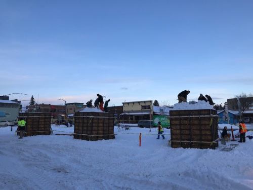 Workers on top of large snow piles in a snowy urban area, with buildings and a clear sky in the background.