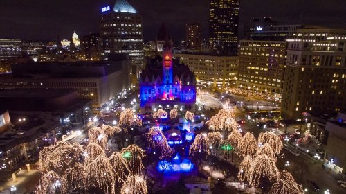 Aerial view of a city square at night, featuring a historic building illuminated with colorful lights and decorated trees.