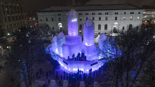 A large illuminated ice castle with towers, surrounded by trees and a crowd, set against a snowy night sky.