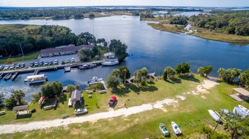 Aerial view of a marina with boats, grassy areas, and a winding river surrounded by trees and buildings.