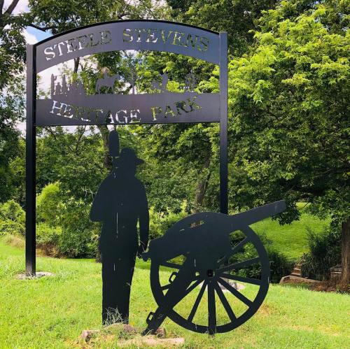 Sign for Steele Stevens Heritage Park featuring a silhouette of a person with a cannon, surrounded by greenery.