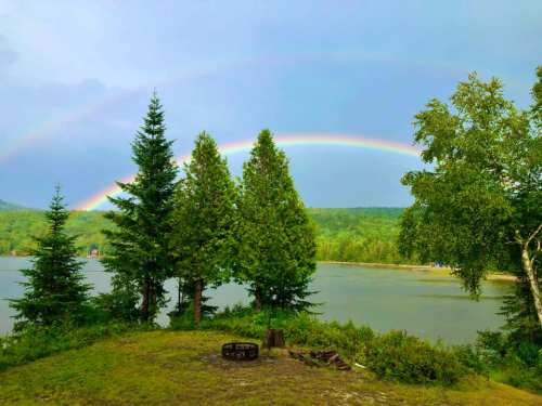 A serene landscape featuring a rainbow over a lake, framed by lush green trees and a fire pit in the foreground.
