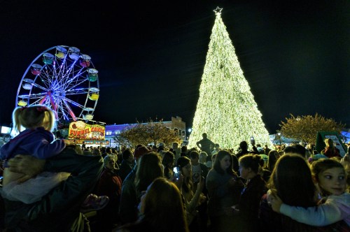A festive crowd gathers around a large illuminated Christmas tree and a colorful Ferris wheel at night.