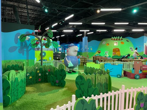 Colorful indoor play area with cartoon characters, green hills, and playful decorations, featuring a fenced garden.