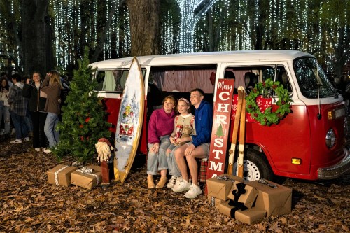 A family poses by a festive red van decorated for Christmas, surrounded by lights, gifts, and holiday decor.