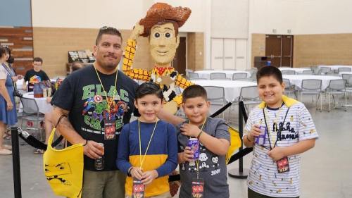 A man and three boys pose with a Woody figure from Toy Story at an event, holding drinks and wearing lanyards.