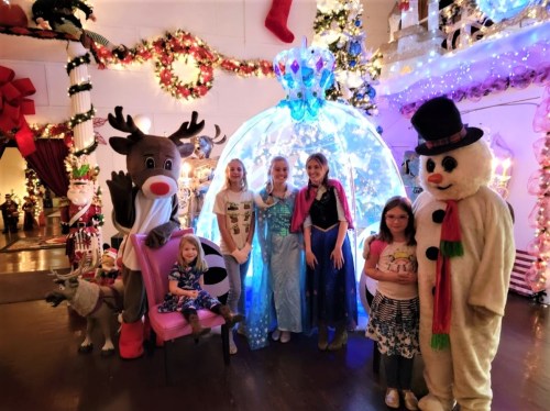 A festive scene with children in costumes, a reindeer, a snowman, and a glowing igloo, surrounded by holiday decorations.