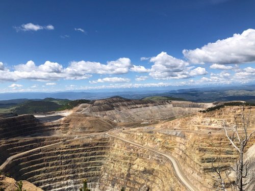 A vast open-pit mine with layered earth and rock, under a blue sky with scattered clouds and distant mountains.