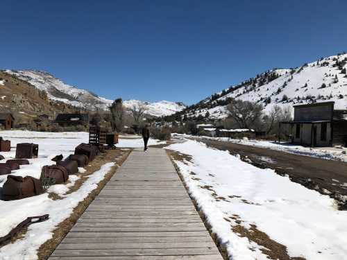 A person walks along a wooden path in a snowy landscape, surrounded by mountains and remnants of an old town.