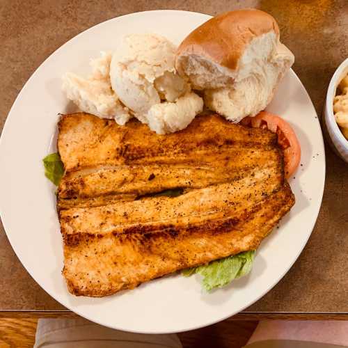 A plate with grilled fish fillet, mashed potatoes, a roll, and a slice of tomato on lettuce.