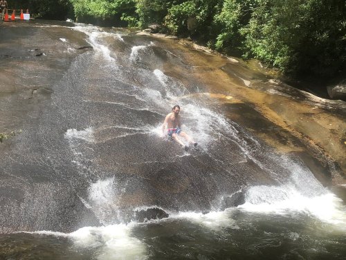 A person slides down a smooth rock into a flowing stream, surrounded by lush greenery.