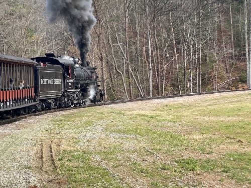 A steam locomotive emits smoke while rounding a curve in a wooded area, with a vintage passenger car attached.