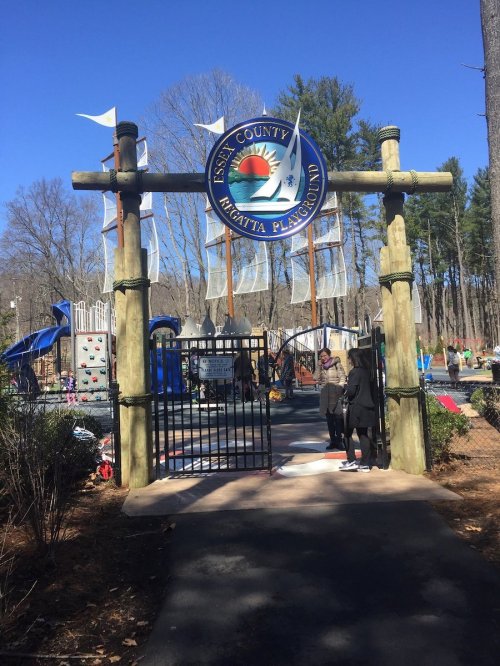 Entrance to a playground with a sign reading "Fisher County Kiwanis Playground," surrounded by trees and blue skies.
