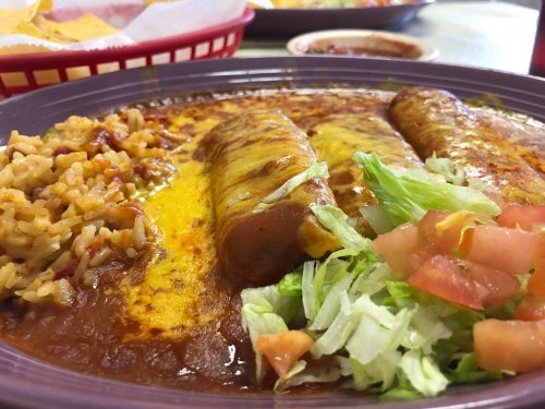 A plate of enchiladas topped with cheese, served with rice, lettuce, and diced tomatoes.