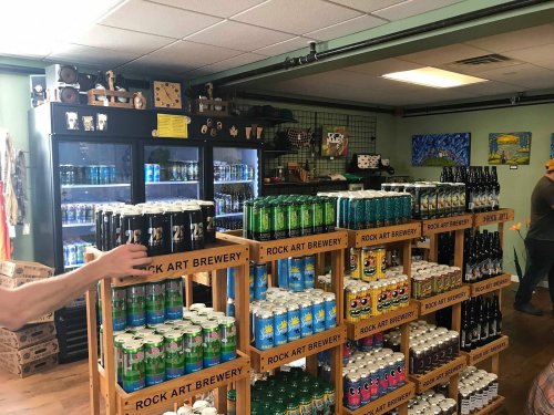 Interior of a brewery store with shelves of colorful canned beverages and a cooler in the background.